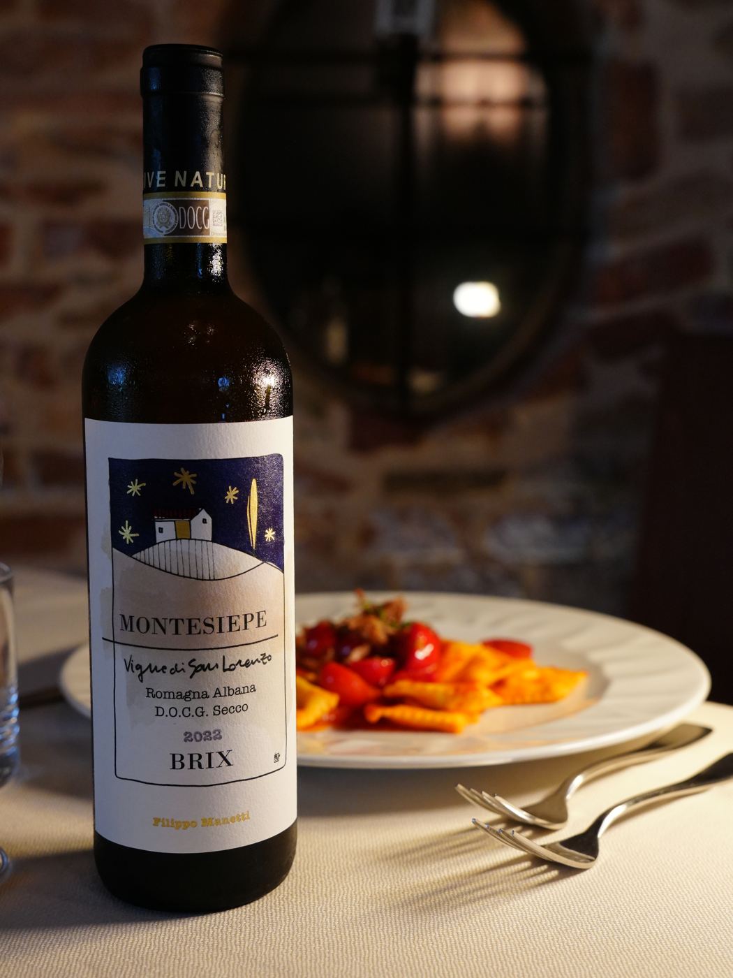 Una bottiglia di Montesiepe Brix di Vigne di San Lorenzo sul tavolo apparecchiato, davanti al muro in mattoni
